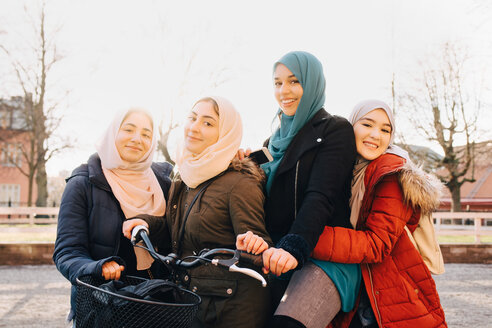 Porträt von glücklichen multiethnischen muslimischen Freundinnen mit Fahrrad in der Stadt gegen den Himmel - MASF00426
