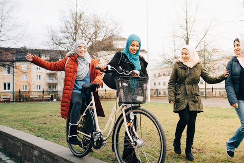 Fröhliche multiethnische Freundinnen gehen mit dem Fahrrad auf einer Wiese in der Stadt spazieren - MASF00429