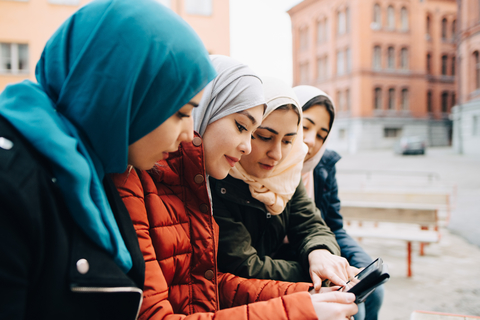 Freundinnen mit Hidschabs sitzen in der Stadt und teilen sich ein digitales Tablet, lizenzfreies Stockfoto