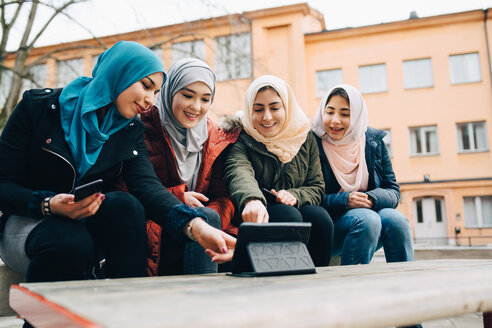 Glückliche muslimische Freundinnen sitzen mit digitalem Tablet auf einer Bank in der Stadt - MASF00435