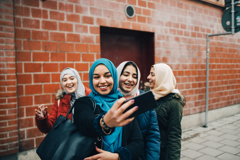 Glückliche junge muslimische Frau, die ein Selfie mit Freundinnen auf dem Bürgersteig vor einem Gebäude in der Stadt macht, lizenzfreies Stockfoto