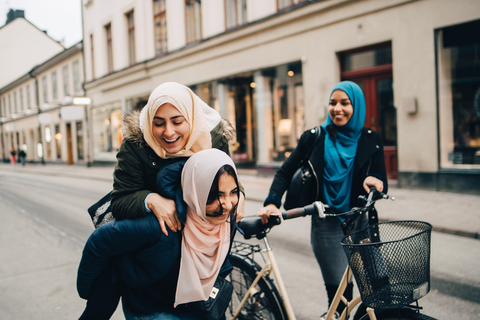 Cheerful teenage girl giving young woman piggyback by friend walking with bicycle on street in city stock photo