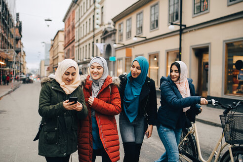 Multiethnische muslimische Freunde gehen mit Fahrrad und Mobiltelefon auf einer Straße in der Stadt spazieren - MASF00447