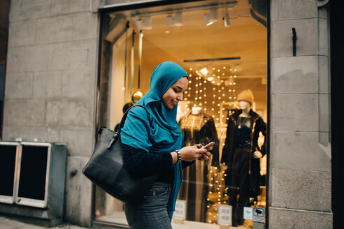 Lächelnde junge Frau, die ein Smartphone benutzt und an einem Schaufenster in der Stadt vorbeigeht - MASF00448