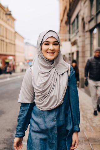Porträt einer lächelnden jungen muslimischen Frau mit Hidschab, die auf dem Bürgersteig in der Stadt spazieren geht, lizenzfreies Stockfoto