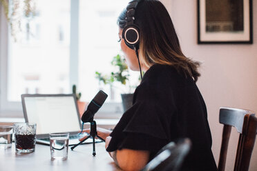 Side view of female podcaster wearing headphones sitting at table with microphone at home - MASF00456