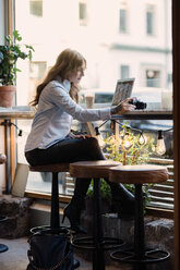 Junge Frau hält Kamera in voller Länge, während sie mit Laptop in einem Café sitzt - MASF00476