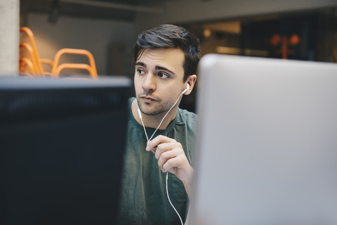 Nachdenklicher Computerprogrammierer trägt In-Ear-Kopfhörer im Büro, lizenzfreies Stockfoto
