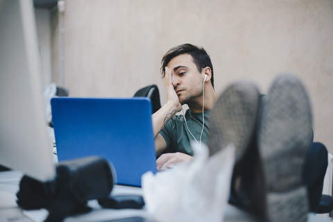 Müder Computerprogrammierer, der einen Laptop benutzt, während er mit hochgelegten Füßen am Schreibtisch im Büro sitzt, lizenzfreies Stockfoto