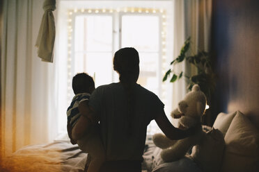 Woman keeping teddy bear on bed while carrying son at bedroom - MASF00494