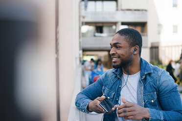 Smiling young man listening to headphones while standing on footbridge in city - MASF00506