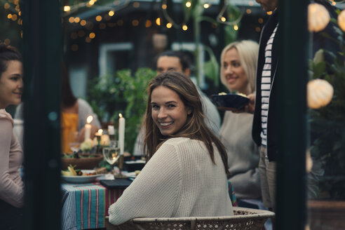 Lächelnde junge Frau schaut zurück, während sie mit Freunden in einem gläsernen Wintergarten im Hinterhof sitzt - MASF00509