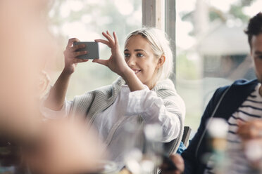 Smiling young woman photographing while sitting by male friend against window - MASF00512