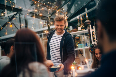 Smiling young man offering fruits to friends in glass cabin - MASF00523