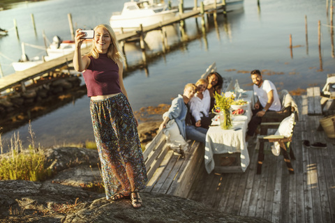 Frau nimmt Selfie durch Smartphone mit Freunden sitzen im Hintergrund am Hafen, lizenzfreies Stockfoto