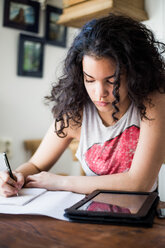 Teenage girl studying at table while writing in book by digital tablet - MASF00570