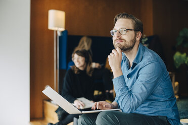 Thoughtful businessman looking away while sitting with laptop by female colleague at office cafeteria - MASF00577