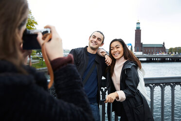 Woman photographing smiling friends standing on bridge by railing in city against clear sky - MASF00586