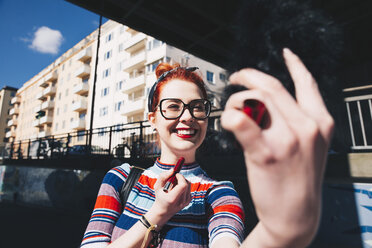 Young woman looking at mobile phone while applying lipstick in city - MASF00604