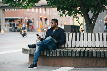 Junger Mann benutzt sein Smartphone, während er mit gekreuzten Beinen auf einer Bank in der Stadt sitzt - MASF00625