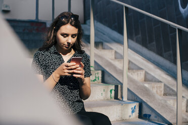 Junge Frau benutzt ihr Smartphone, während sie an einem sonnigen Tag auf einer Treppe sitzt - MASF00631