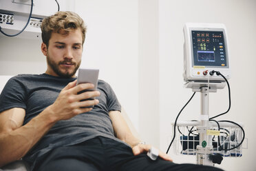 Junger männlicher Patient, der sein Smartphone benutzt, während er auf dem Bett liegt, während eines medizinischen Tests im Krankenhaus - MASF00636
