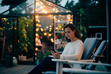 Young woman using mobile phone while sitting on lounge chair against glass cabin - MASF00666