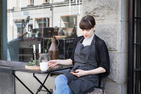 Junge Frau, die ihr Smartphone benutzt, während sie mit einer Kaffeetasse am Tisch vor einem Geschäft sitzt - MASF00686