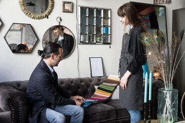 Young female owner looking at mature male customer choosing from fabric swatch while sitting on sofa at store - MASF00693