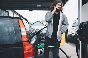 Junger Mann spricht beim Tanken an einer Tankstelle mit seinem Handy - MASF00696