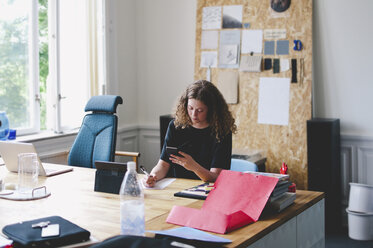 Junge Geschäftsfrau, die ein Mobiltelefon benutzt, während sie am Schreibtisch in einem kreativen Büro auf Papier schreibt - MASF00702