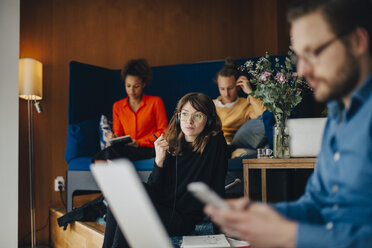 Young businesswoman listening to headphones while sitting amidst colleagues at office in cafeteria - MASF00710