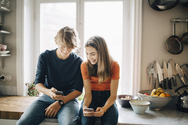 Teenager, der einer Freundin sein Handy zeigt, während er am Küchentisch sitzt - MASF00745