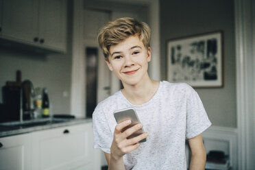 Portrait of smiling teenage boy holding smart phone while sitting in kitchen at home - MASF00748