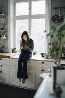 Full length of smiling girl using mobile phone while sitting on kitchen counter - MASF00749