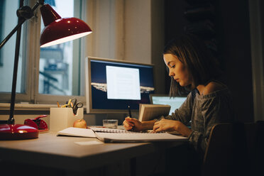 Side view of girl writing while using computer at illuminated desk - MASF00777