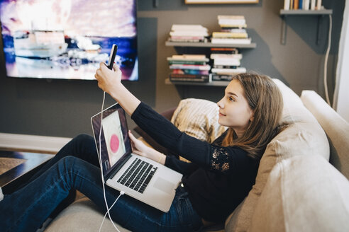 Seitenansicht eines Mädchens, das ein Selfie mit dem Mobiltelefon macht, während es einen Laptop auf dem Sofa benutzt - MASF00790