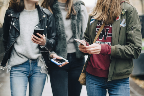 Mittelteil von Freundinnen, die beim Spazierengehen in der Stadt ein Mobiltelefon benutzen, lizenzfreies Stockfoto