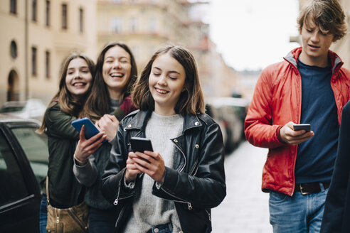 Lächelnde Freunde benutzen Mobiltelefone, während sie auf dem Bürgersteig in der Stadt spazieren gehen - MASF00813