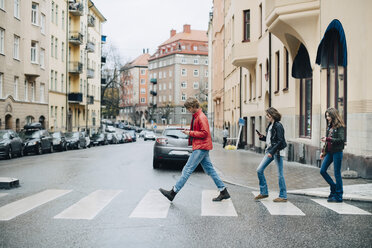 Freunde in voller Länge, die beim Überqueren der Straße in der Stadt ein Mobiltelefon benutzen - MASF00815