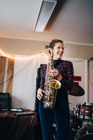 Glückliche junge Musikerin spielt Saxophon im Proberaum, lizenzfreies Stockfoto