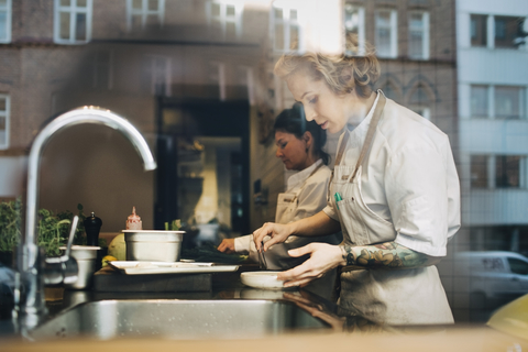 Seitenansicht von weiblichen Köchen, die am Tresen in einer Restaurantküche Essen zubereiten, vom Fenster aus gesehen, lizenzfreies Stockfoto