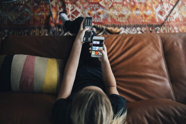 Blick von oben auf ein Teenager-Mädchen, das eine Handy-App und eine Fernbedienung benutzt, während es auf dem Sofa sitzt und zu Hause fernsieht - MASF00900