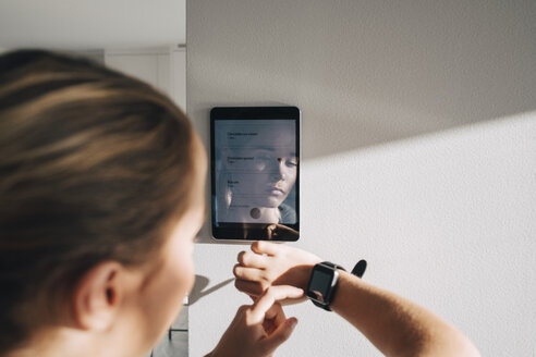 Teenage girl adjusting smart watch with digital tablet mounted on white wall at home - MASF00908