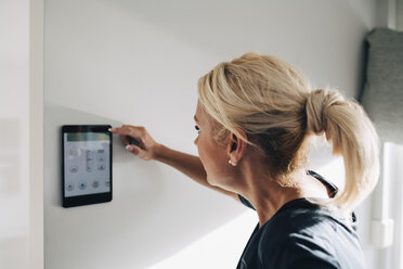 Side view of blond woman adjusting thermostat using digital tablet mounted on white wall at home - MASF00913