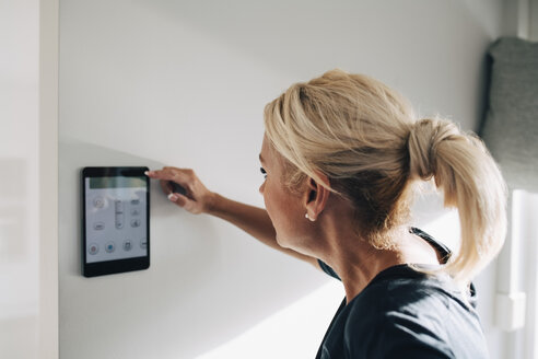 Seitenansicht einer blonden Frau, die den Thermostat mithilfe eines digitalen Tablets an einer weißen Wand zu Hause einstellt - MASF00913