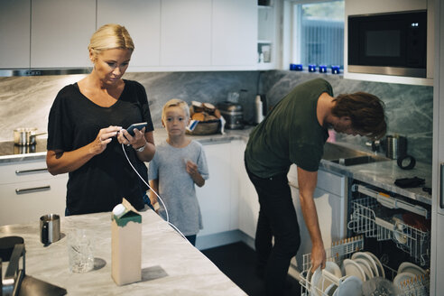 Junge steht neben seiner Mutter, die ein Mobiltelefon benutzt, und seinem Vater, der in der Küche Teller im Geschirrspüler reinigt - MASF00916