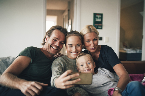 Lächelnde Familie, die ein Selfie mit ihrem Smartphone macht, während sie zu Hause auf dem Sofa sitzt, lizenzfreies Stockfoto