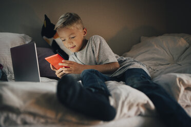 Junge in voller Länge, der ein Handy benutzt und sich mit einem Laptop auf dem Bett zu Hause zurücklehnt - MASF00933