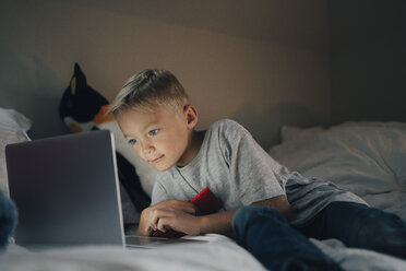 Junge, der einen Laptop benutzt und sich mit seinem Mobiltelefon auf dem Bett zurücklehnt - MASF00936